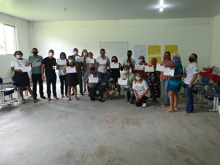 DESCRIÇÃO: foto colorida de 21 pessoas posando para a foto, cada uma com um certificado na mão. Ao fundo há a parede branca, com um quadro branco e alguns cartazes em cartolina amarela grudados na parede.